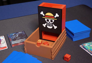 One Piece Dice Tower opened up with red and gold dice in the tray and next to it while sitting on a black playmat next to a Belo Betty deck from OPTCG