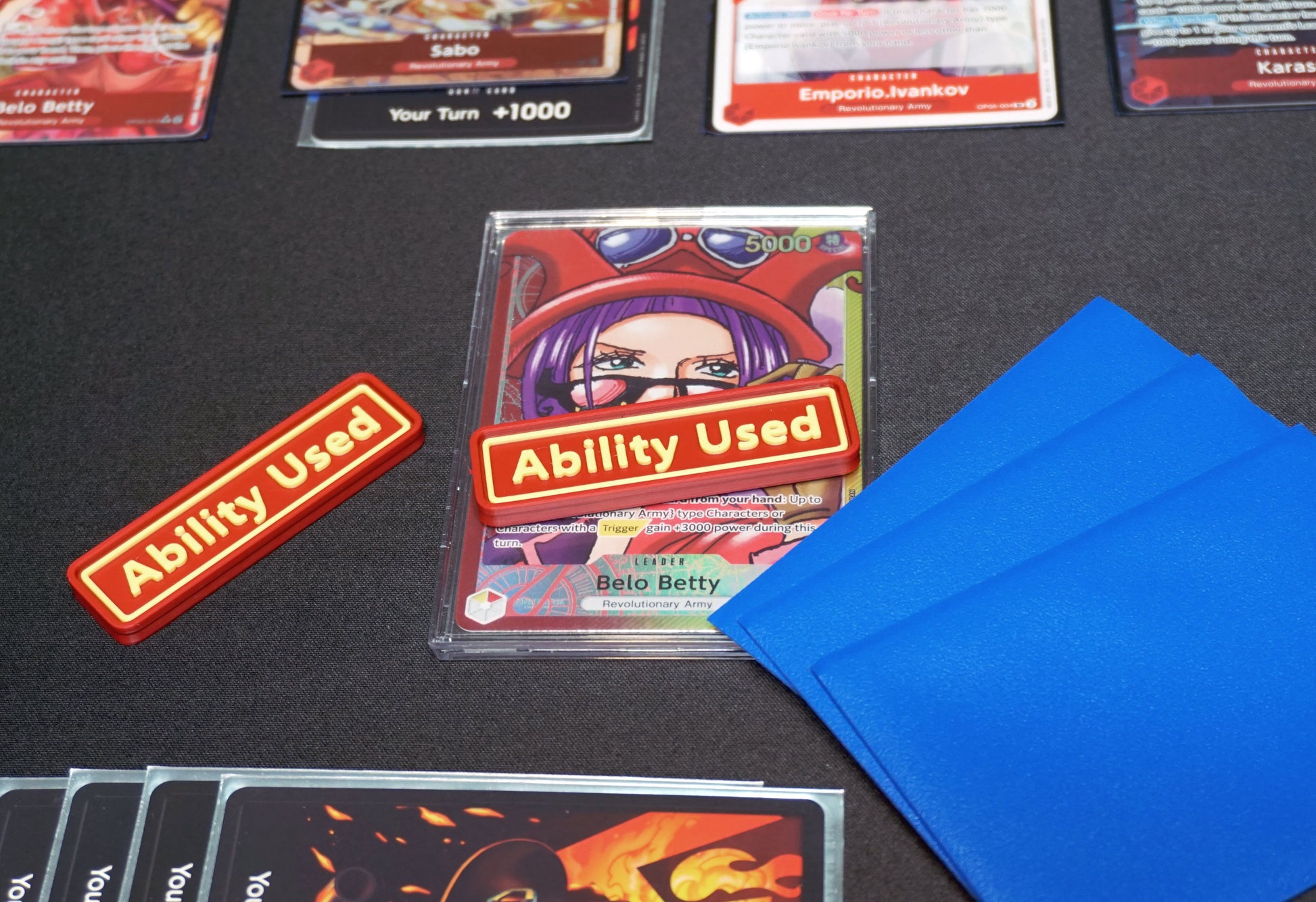 A Red and Yellow Ability User Marker sits on top of the Belo Betty leader alt art card with the hand face down next to it, all on top of a black playmat