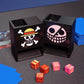 One Piece Mini Dice Towers of the Strawhats and Donquixote jolly rogers on a black playmat with red/gold and pink/gold dice next to a Belo Betty OPTCG deck