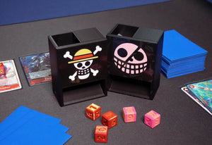 One Piece Mini Dice Towers of the Strawhats and Donquixote jolly rogers on a black playmat with red/gold and pink/gold dice next to a Belo Betty OPTCG deck
