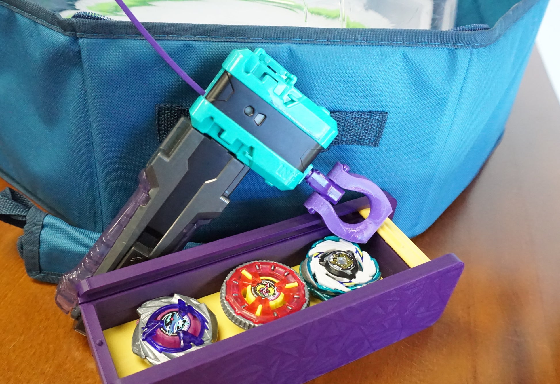 The Slip Grip in purple attached to the teal rip cord launcher next to the purple garage door style deckbox with 3 beyblades inside