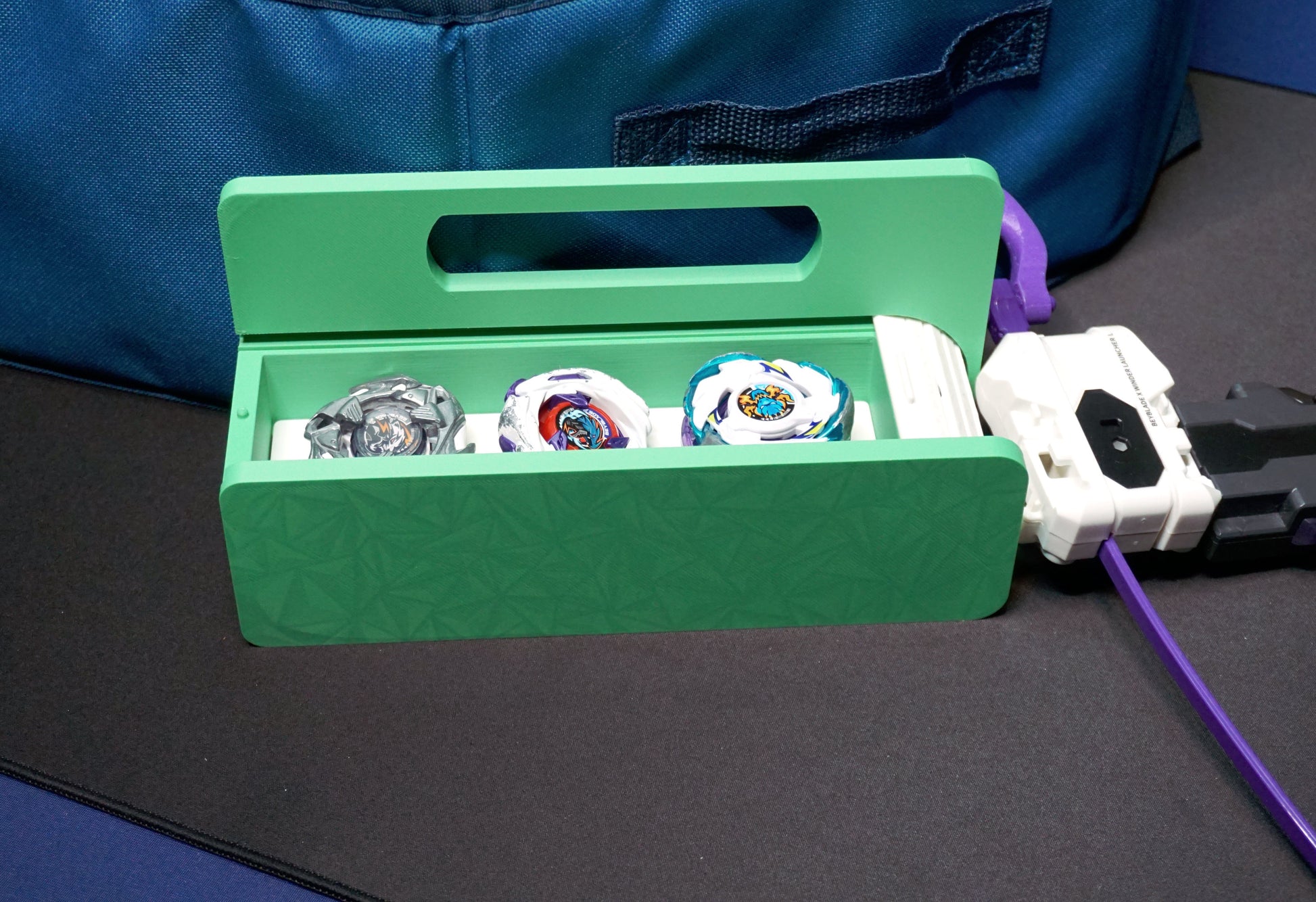 The Garage Door style deck box for Beyblade X in green for the base with a white door and tray. With Weiss Tiger, Meteor Dragoon, Dran Blast inside the case. Next to it is a Takara Tomy stadium in a case with a white launcher and purple rip cord with a slip grip attachment and handle
