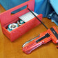 A red base with silver door and tray Garage Door style deckbox sitting on a wooden table. Next to it is a Takara Tomy stadium in a case and a red ripcord launcher with red handle and cherry red slip grip attachment. Inside the deck box is Emperor Blast and Wolf Hunt