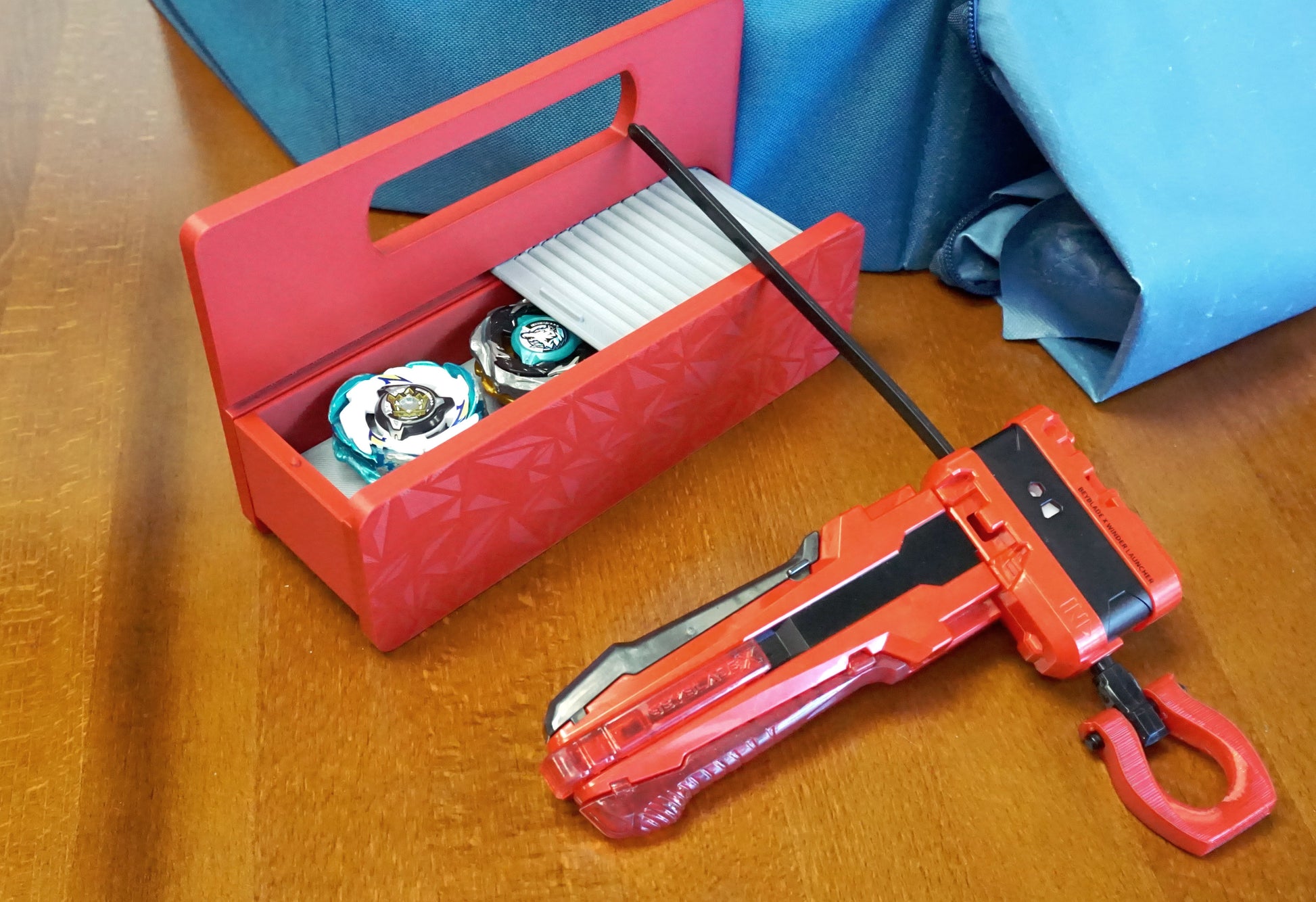 A red base with silver door and tray Garage Door style deckbox sitting on a wooden table. Next to it is a Takara Tomy stadium in a case and a red ripcord launcher with red handle and cherry red slip grip attachment. Inside the deck box is Emperor Blast and Wolf Hunt