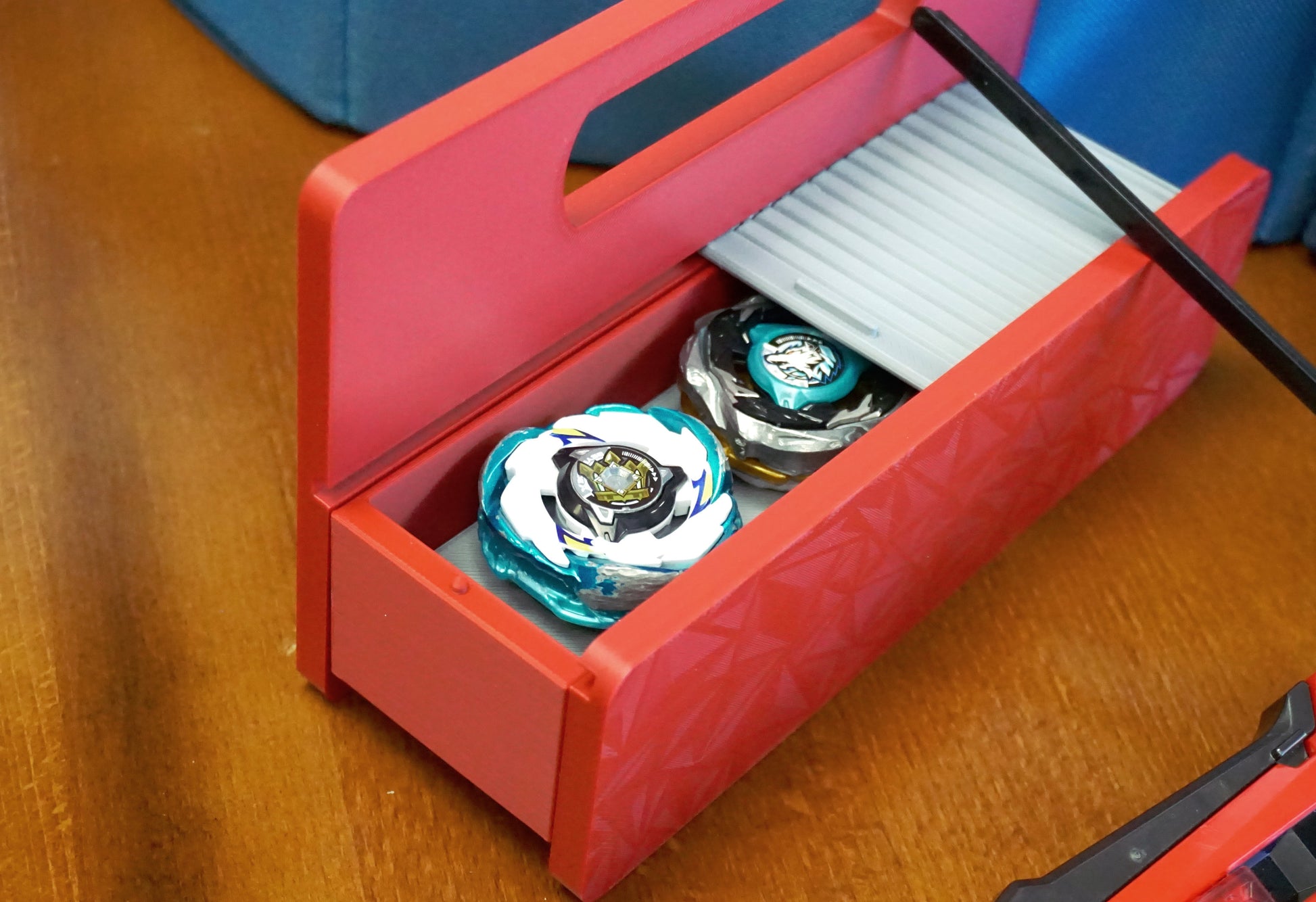 A red base with silver door and tray Garage Door style deckbox sitting on a wooden table. Next to it is a Takara Tomy stadium in a case and a red ripcord launcher with red handle and cherry red slip grip attachment. Inside the deck box is Emperor Blast and Wolf Hunt