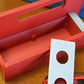 A red base with silver door and tray Garage Door style deckbox sitting on a wooden table. Next to it is a Takara Tomy stadium in a case and a red ripcord launcher with red handle and cherry red slip grip attachment. 