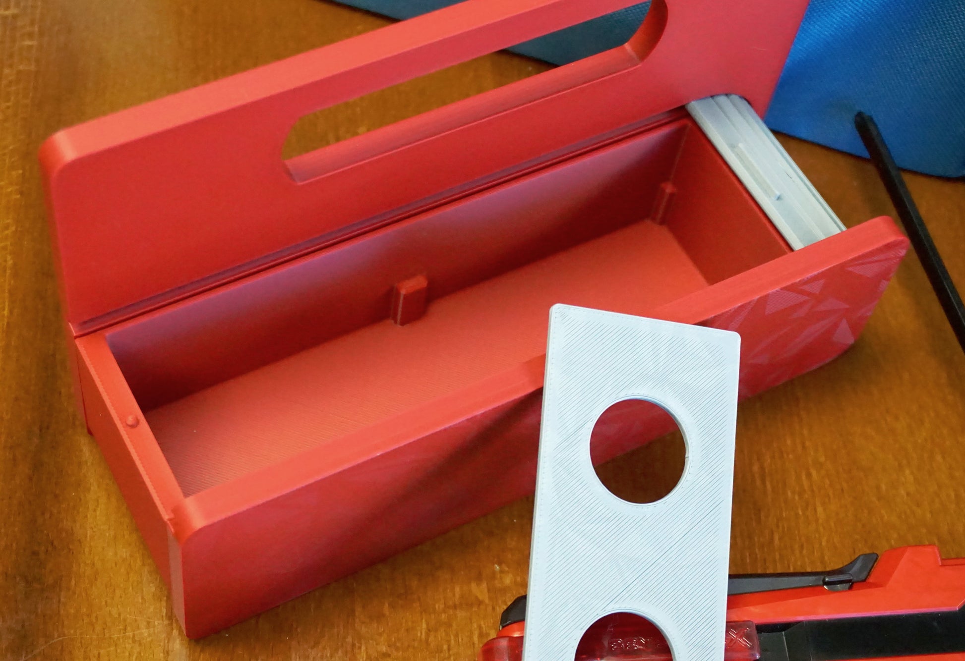 A red base with silver door and tray Garage Door style deckbox sitting on a wooden table. Next to it is a Takara Tomy stadium in a case and a red ripcord launcher with red handle and cherry red slip grip attachment. 
