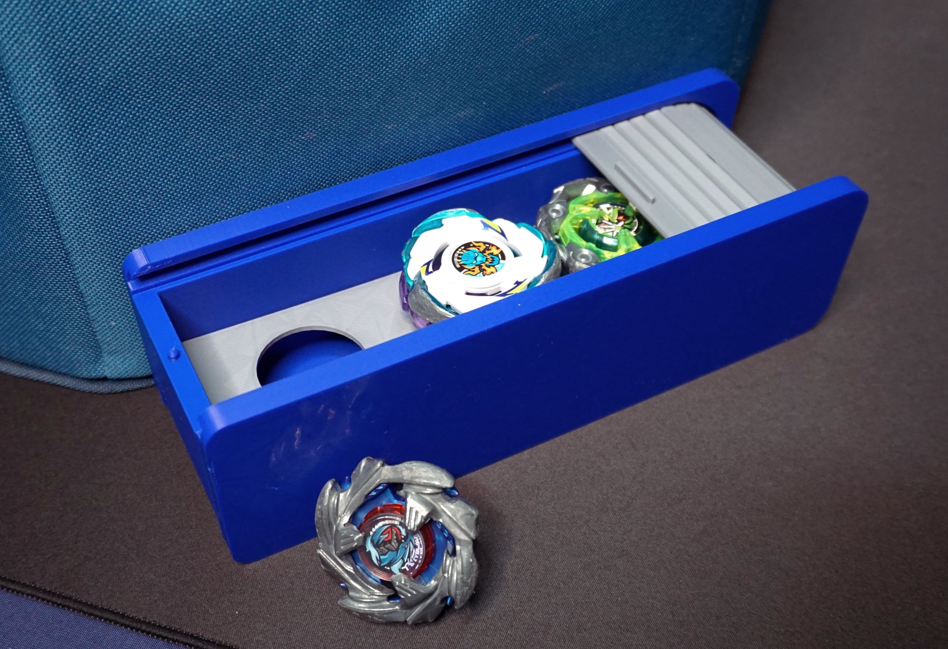 The Beyblade X Garage Door style deckbox from Momo's Models shown in blue with grey color scheme. Next to a carrying case for the Takara Tomy stadium with the Cobalt Dragoon, Pegasus Blast, and Wizard Rod beyblades.