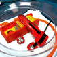 The Beyblade X Garage Door style deckbox from Momo's Models shown in the red and orange color scheme for good luck. Sitting inside the Takara Tomy stadium with a red ripcord launcher that has a red handle and red slip grip.