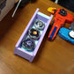 The Beyblade X Garage Door style deckbox from Momo's Models shown in the lavender and glacier color scheme. Sitting on a wood table next to a red and blue string launchers with grips.