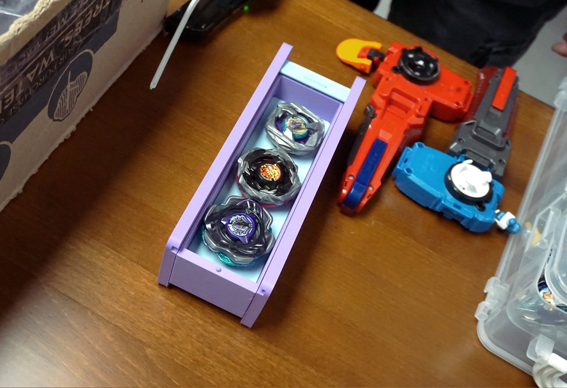 The Beyblade X Garage Door style deckbox from Momo's Models shown in the lavender and glacier color scheme. Sitting on a wood table next to a red and blue string launchers with grips.