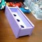 An angled shot of the Beyblade X Garage Door style deckbox from Momo's Models shown in the lavender and glacier color scheme. Sitting on a wood table next to a teal ripcord launcher along with red and blue string launchers with grips.