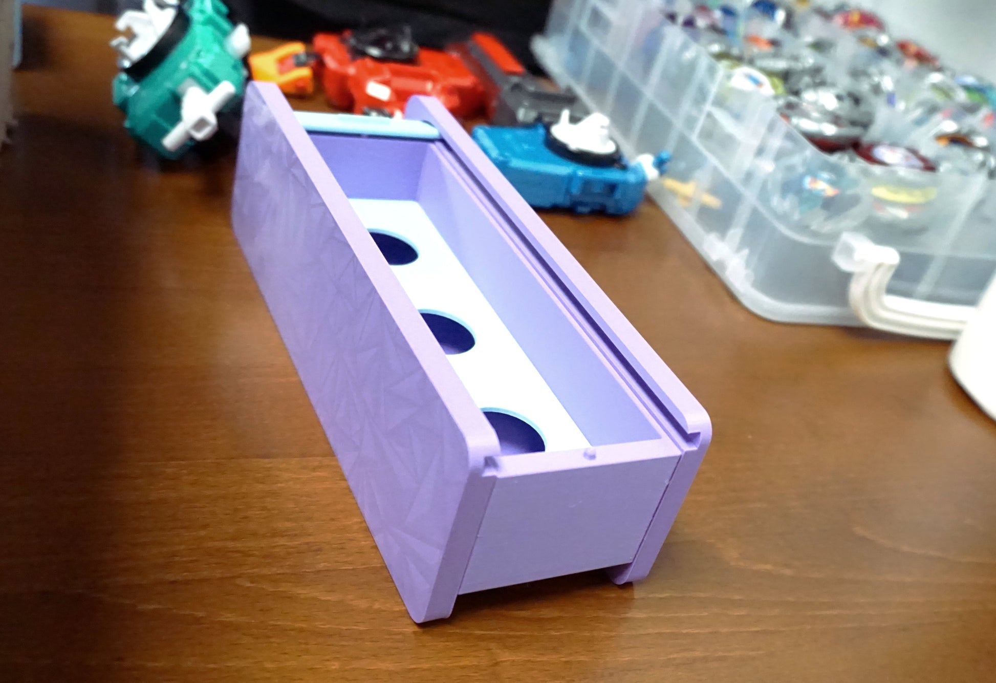 An angled shot of the Beyblade X Garage Door style deckbox from Momo's Models shown in the lavender and glacier color scheme. Sitting on a wood table next to a teal ripcord launcher along with red and blue string launchers with grips.