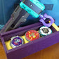 The Beyblade X Garage Door style deckbox from Momo's Models shown in the purple and yellow color scheme. Sitting on a table against a carrying case for the Takara Tomy stadium and a teal ripcord launcher with grip and purple slip grip. Inside of the deckbox is Shark Scale, Clock Mirage, and Pegasus Blast.