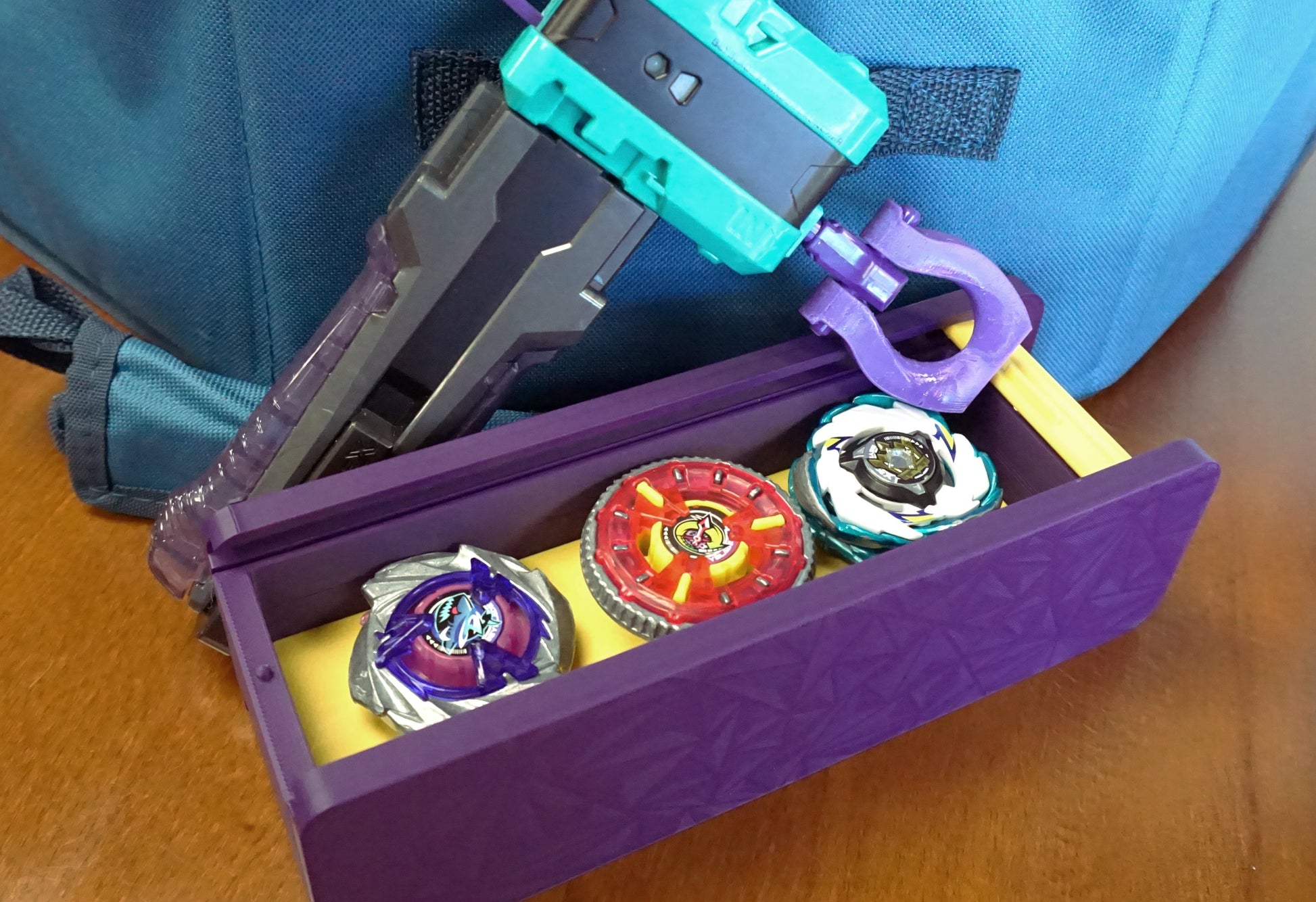 The Beyblade X Garage Door style deckbox from Momo's Models shown in the purple and yellow color scheme. Sitting on a table against a carrying case for the Takara Tomy stadium and a teal ripcord launcher with grip and purple slip grip. Inside of the deckbox is Shark Scale, Clock Mirage, and Pegasus Blast.