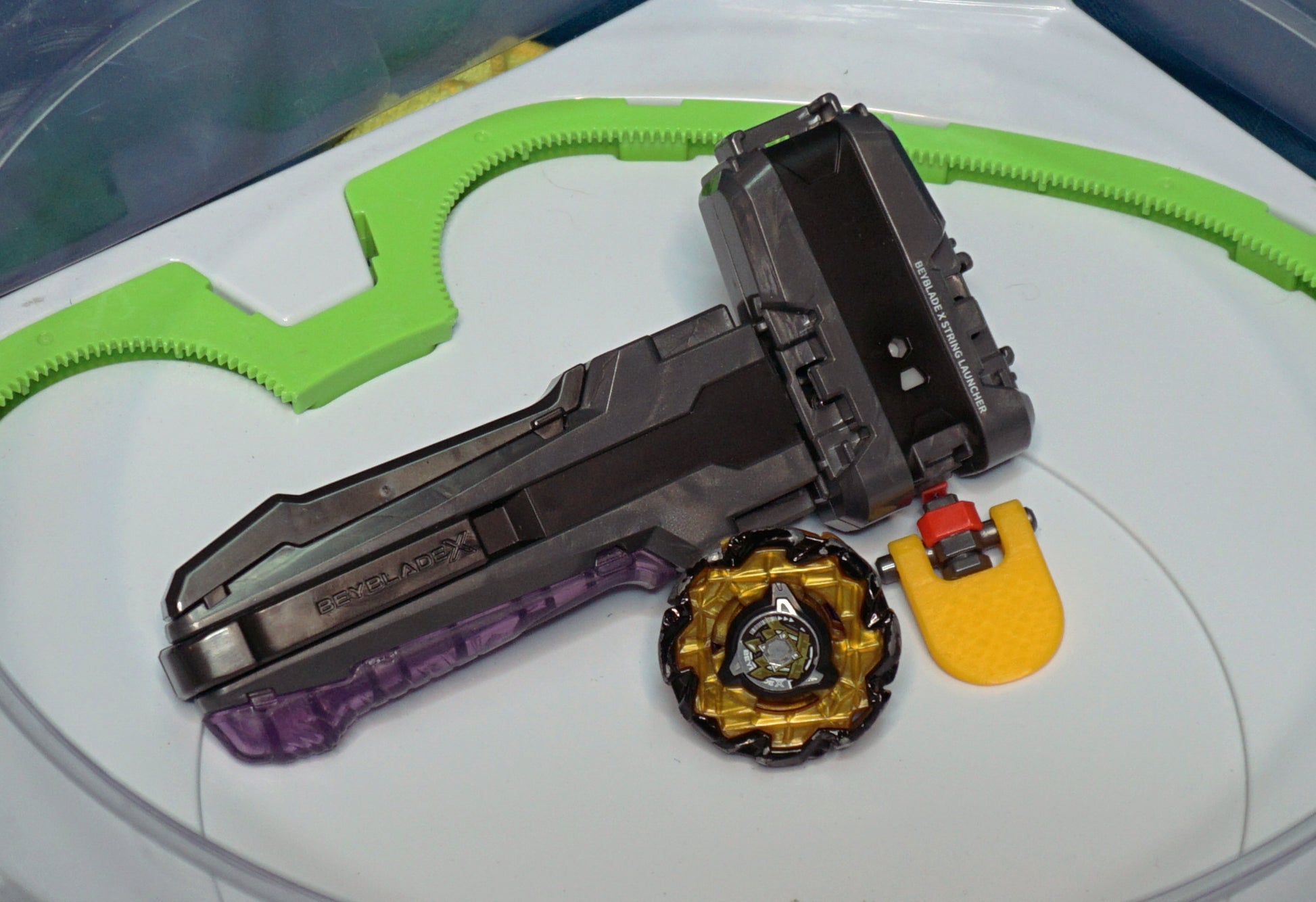 The orange Launcher Tab for Beyblade X on the black/grey string launcher and grip in a Takara Tomy stadium next to Emperor Might blade