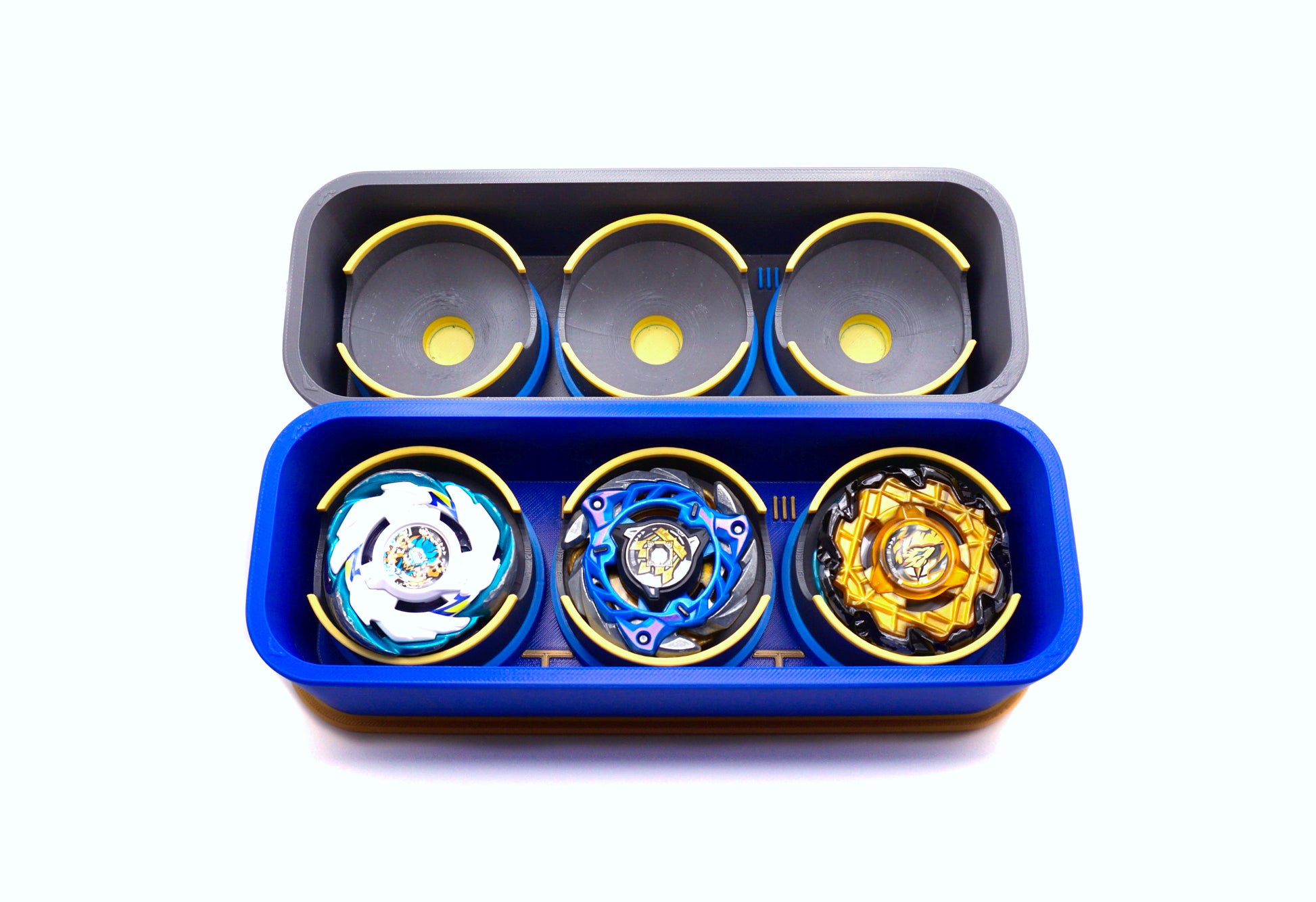 The Beyblade X Deckbox from Momo's Models in 2 different color combinations, black and sky blue for one then blue and gold for the other. Both angled to show the inside cups that hold the beyblades. With Pegasus Blast, Cerberus Flame, and Emperor Might in one of the deckboxes