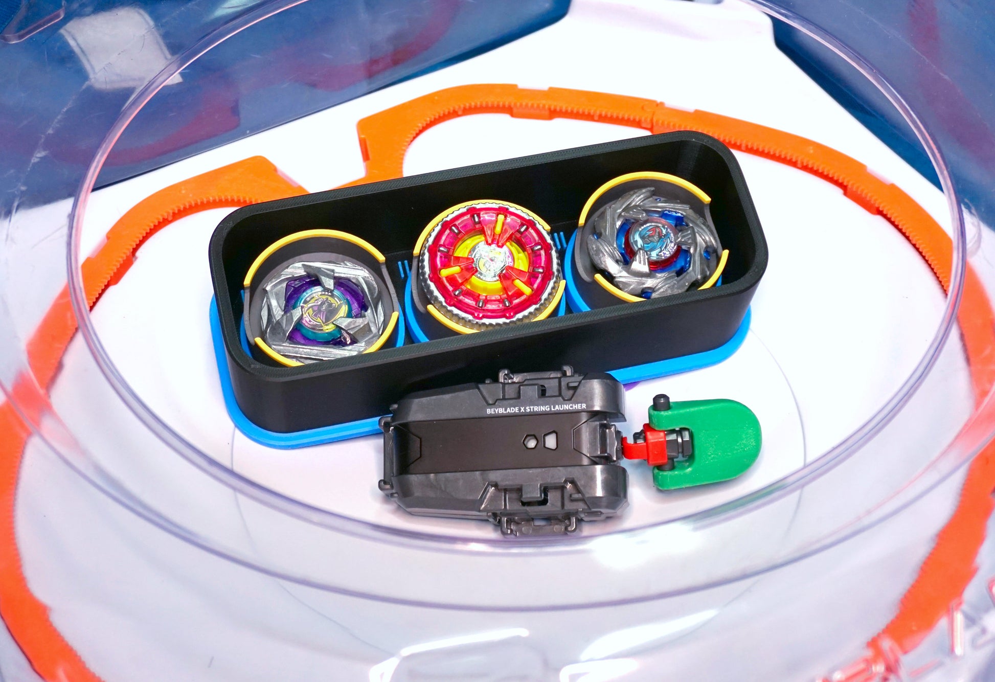The Beyblade X Deckbox from Momo's Models with the Samurai Calibur, Clock Mirage, and Cobalt Dragoon beyblades inside the Takara Tomy Stadium next to a grey string launcher with a green thumb tab