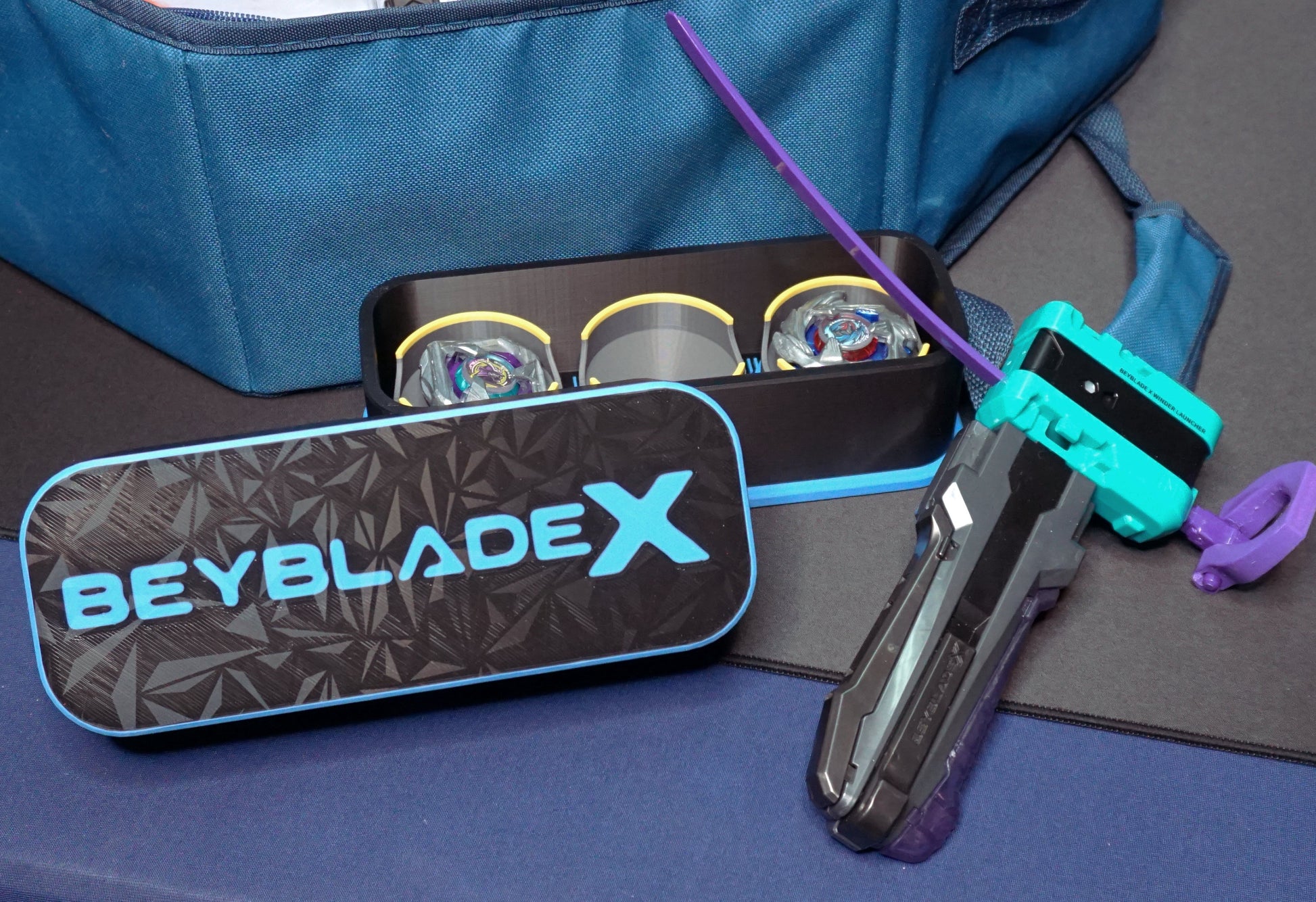 The Beyblade X Deckbox from Momo's Models shown in Black and Sky Blue with the Samurai Calibur and Cobalt Dragoon in the case. Next to a Takara Tomy stadium and teal launcher with a Slip Grip