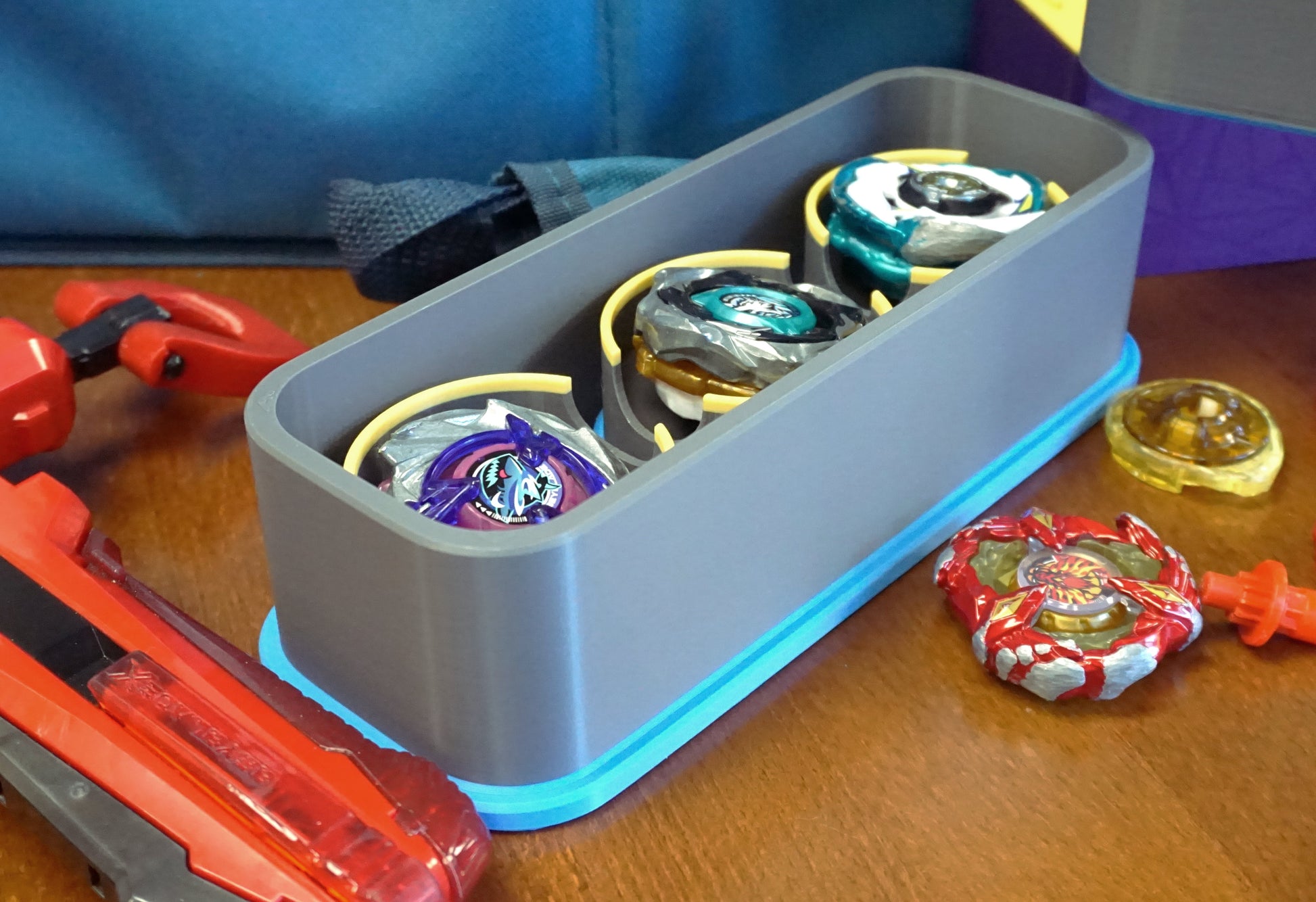 The Beyblade X Deckbox from Momo's Models shown in the Grey and Sky Blue color combination. With a red ripcord launcher and slip grip next to it, along with a few beyblade parts. All on a wooden table next to a Takara Tomy stadium