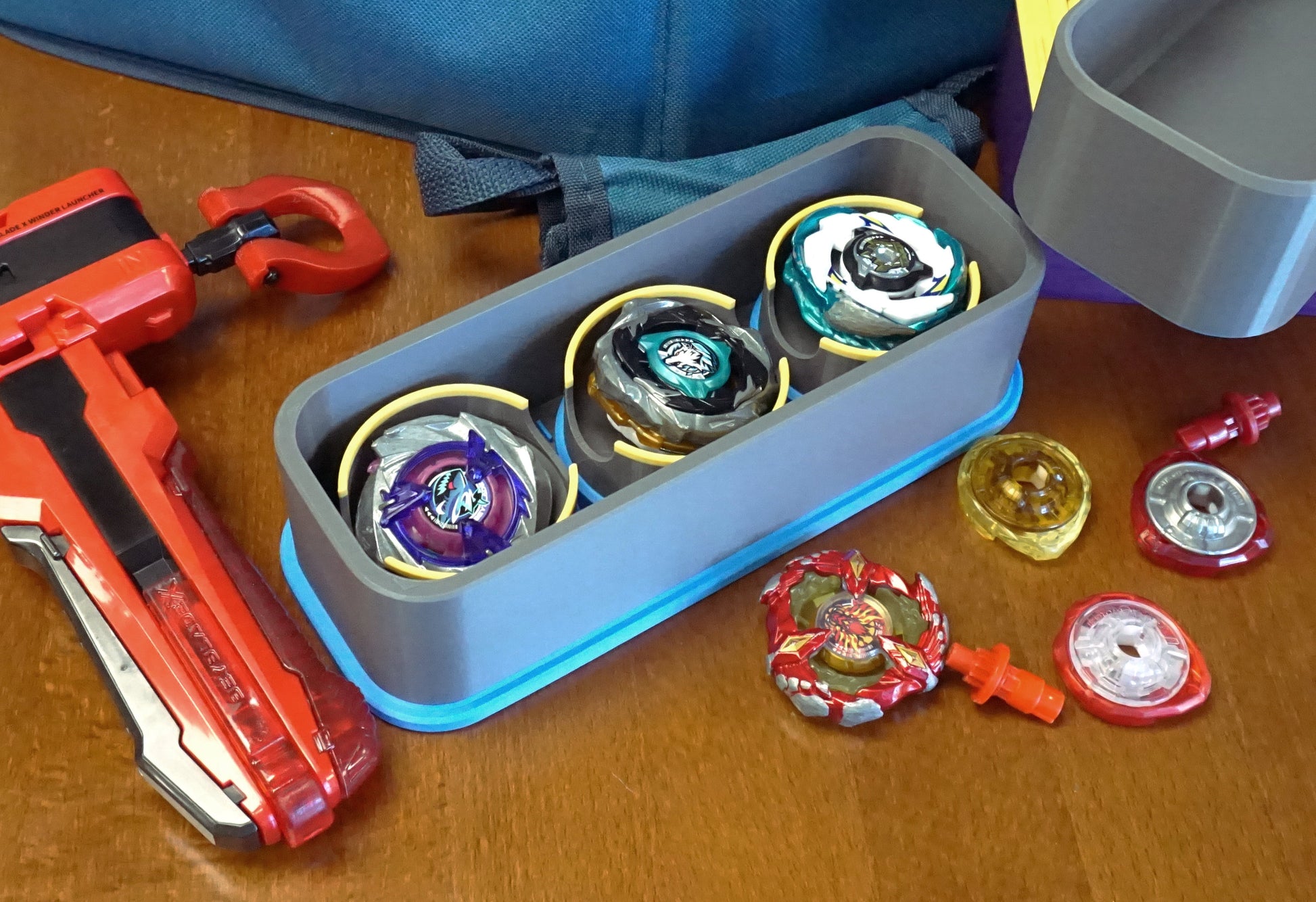 A top down view of the Beyblade X Deckbox from Momo's Models shown in the Grey and Sky Blue color combination. With a red ripcord launcher and slip grip next to it, along with a few beyblade parts. All on a wooden table next to a Takara Tomy stadium
