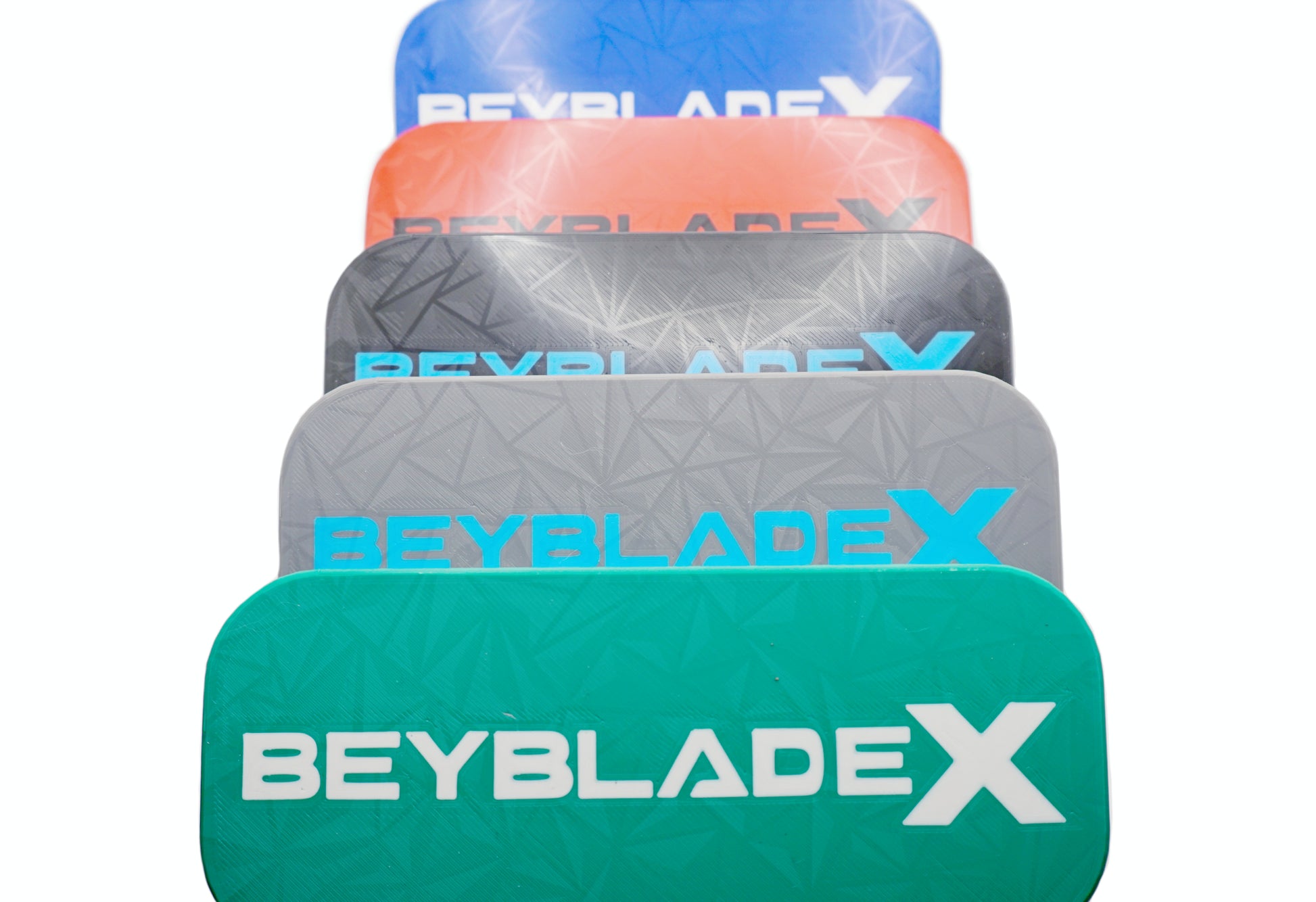 A stack of five Beyblade Pocket deck boxes in shades of green, black, red, gray, and blue. Each deck box features a geometric pattern and the text "BEYBLADE X" in white, black, or sky blue.