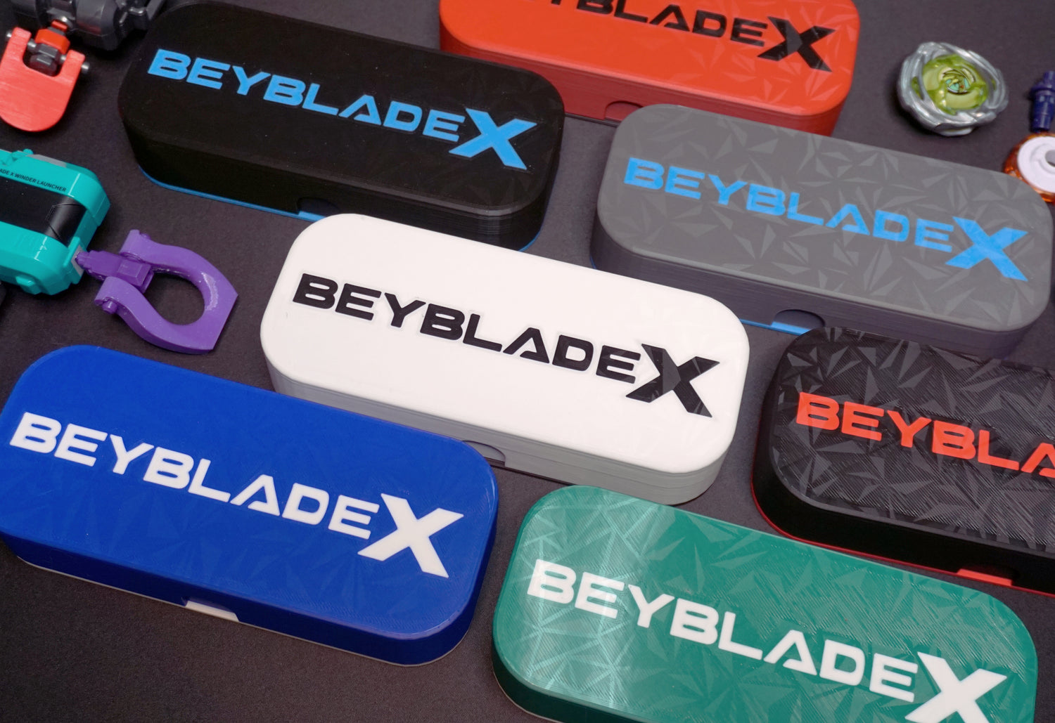 A collection of Beyblade X deck boxes or storage cases in various colors, including black, white, blue, red, gray, and green. Each case features the Beyblade X logo in contrasting colors. Accessories are scattered around the cases.