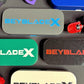 An assortment of rectangular deck cases with the text "BEYBLADE X" in various colors, including gray with blue text, green with white text, and black with red text. Other toy parts are also visible.