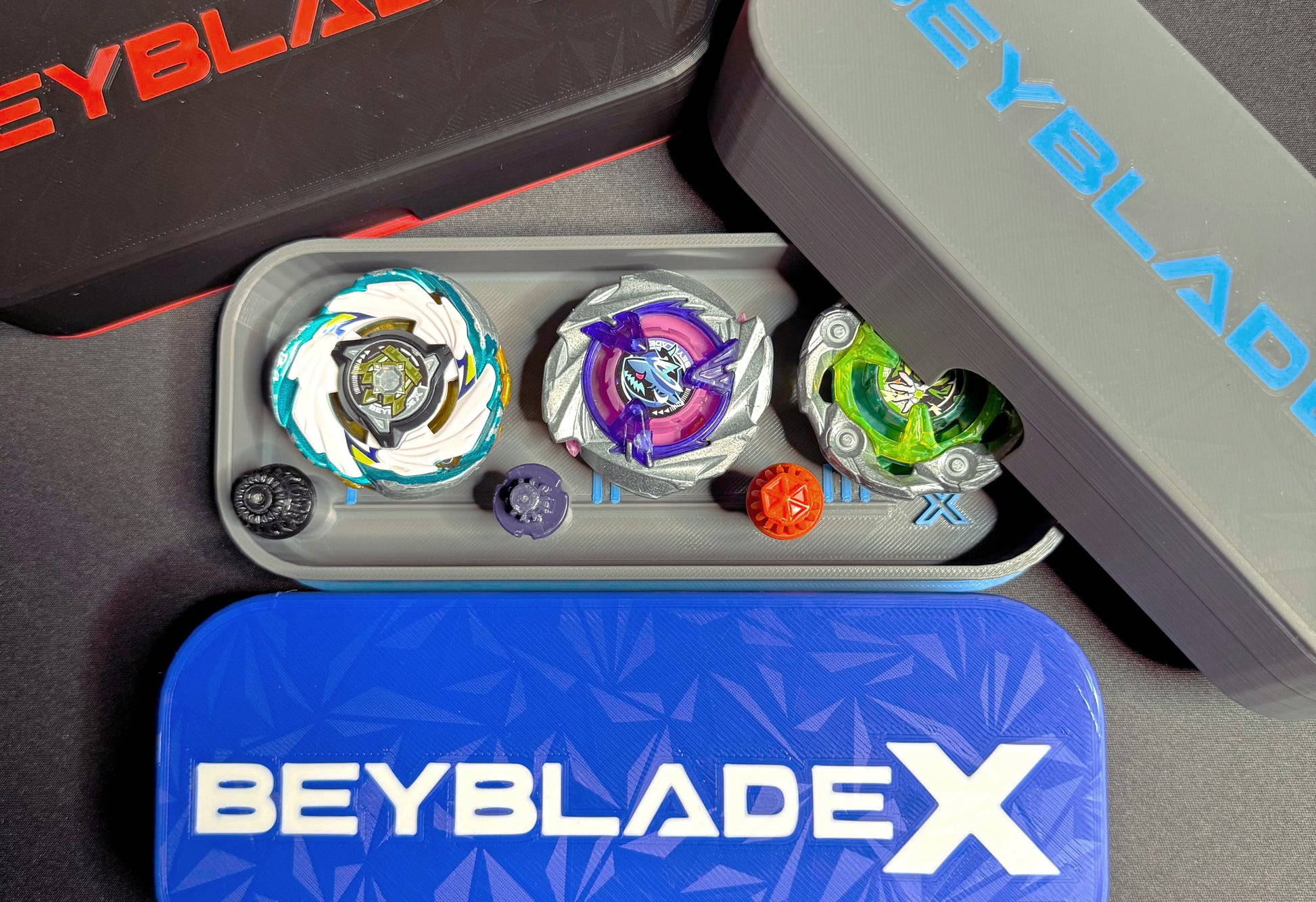 A collection of Beyblade X tops and storage cases. The tops feature various colors and designs, including green, purple, and white. The cases are black, gray, and blue, with the Beyblade X logo.