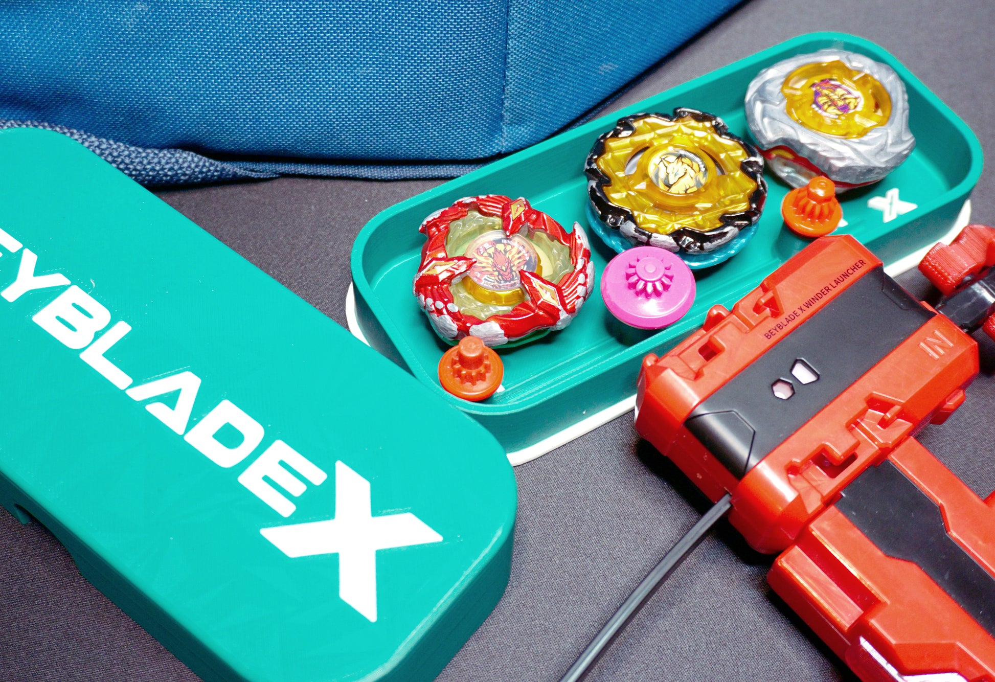 A green container with the text "BEYBLADE X" in white, next to a green tray holding several colorful spinning tops and a red launcher. The tops are various colors, including red, yellow, and silver.