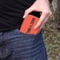 An red Pocket Deck Box with the word "BEYBLADE X" in black is partially visible in a blue jeans pocket. The person is wearing a black t-shirt. The background is blurred.