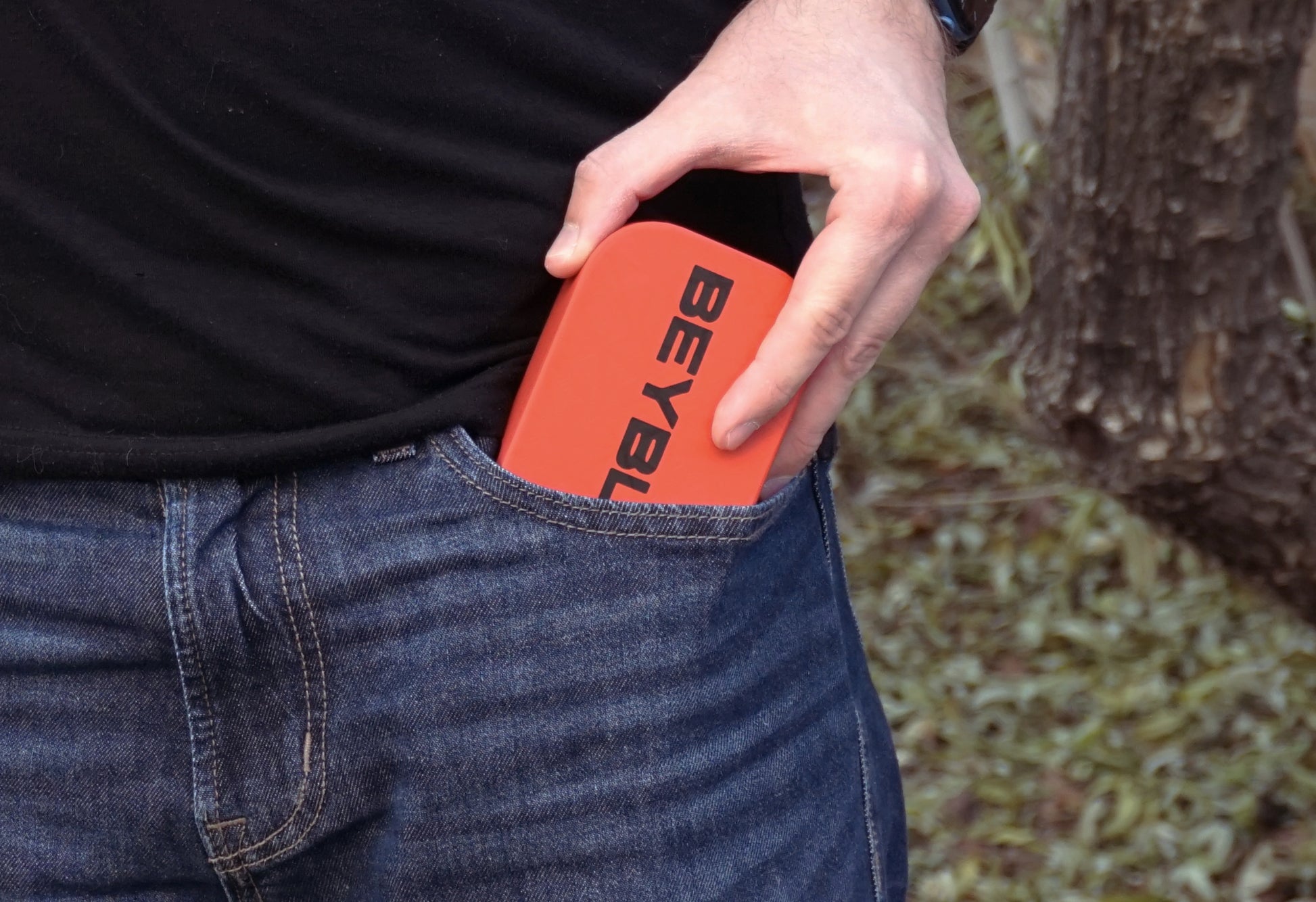 An red Pocket Deck Box with the word "BEYBLADE X" in black is partially visible in a blue jeans pocket. The person is wearing a black t-shirt. The background is blurred.