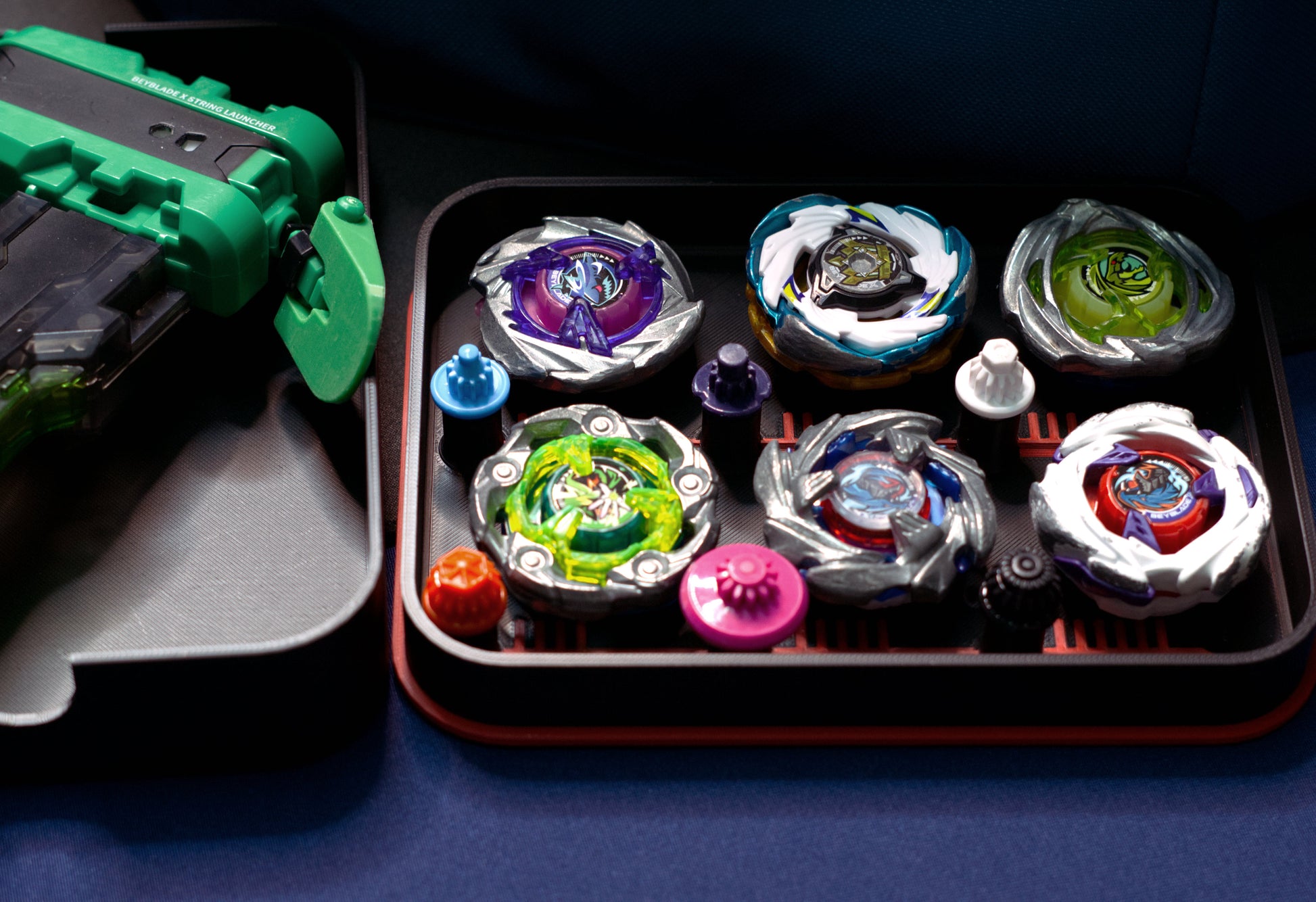 The Pocket Deck Box that holds 6 beys for Beyblade X shown in the black and red color combination. Showcasing Shark Scale, Meteor Dragoon, Wizard Rod, Emperor Pegasus Blast, Cobalt Dragoon, and Hover Wyvern. Sitting on a table with the lid flipped over and holding the green and black string launcher with the green Slip Grip Tab from Momo's Models.