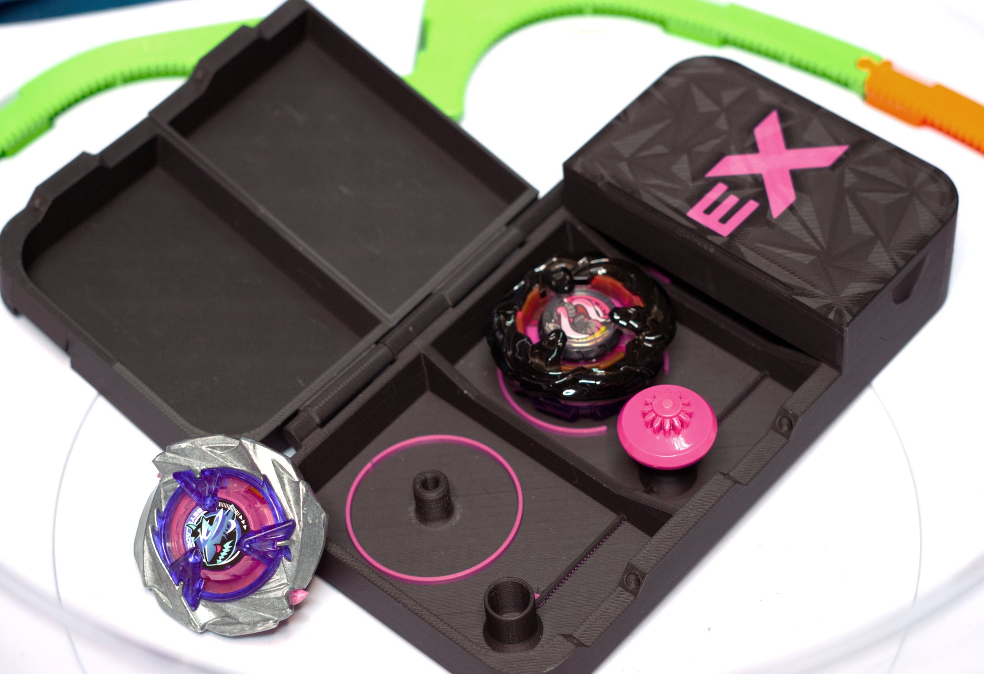 The Beyblade X Pocket Clam Shell Deck Box shown in black and hot pink. Sitting inside a Takara Tomy Extreme Stadium with Shark Scale and Mammoth Tusk beyblades