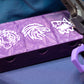 The Beyblade X Pocket Clam Shell Deck Box shown in purple and white with multiple icons on top. Sitting next to a Takara Tomy Extreme Stadium inside a wreath case and the ripcord launcher with handle and purple Slip Grip from Momo's Models