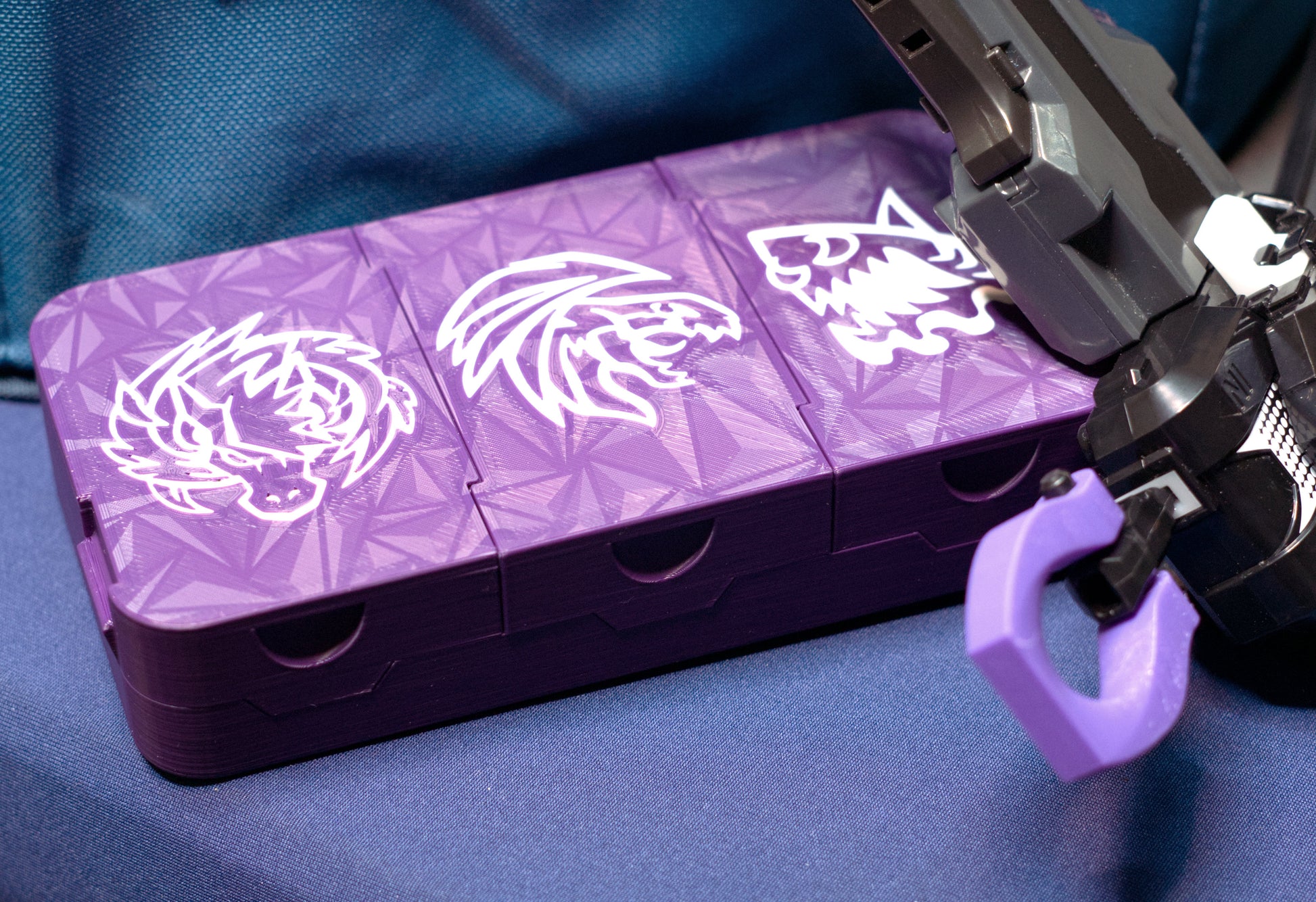 The Beyblade X Pocket Clam Shell Deck Box shown in purple and white with multiple icons on top. Sitting next to a Takara Tomy Extreme Stadium inside a wreath case and the ripcord launcher with handle and purple Slip Grip from Momo's Models