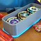 The Beyblade X Deckbox from Momo's Models shown in the Grey and Sky Blue color combination. With a red ripcord launcher and slip grip next to it, along with a few beyblade parts. All on a wooden table next to a Takara Tomy stadium
