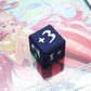 Mini Positive Dice shown in black and white with a +3 on top of a Shirahoshi card from One Piece TCG
