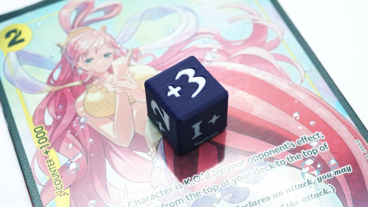 Mini Positive Dice shown in black and white with a +3 on top of a Shirahoshi card from One Piece TCG