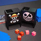 One Piece Mini Dice Towers of the Strawhats and Donquixote jolly rogers on a black playmat with red/gold and pink/gold dice next to a Belo Betty OPTCG deck