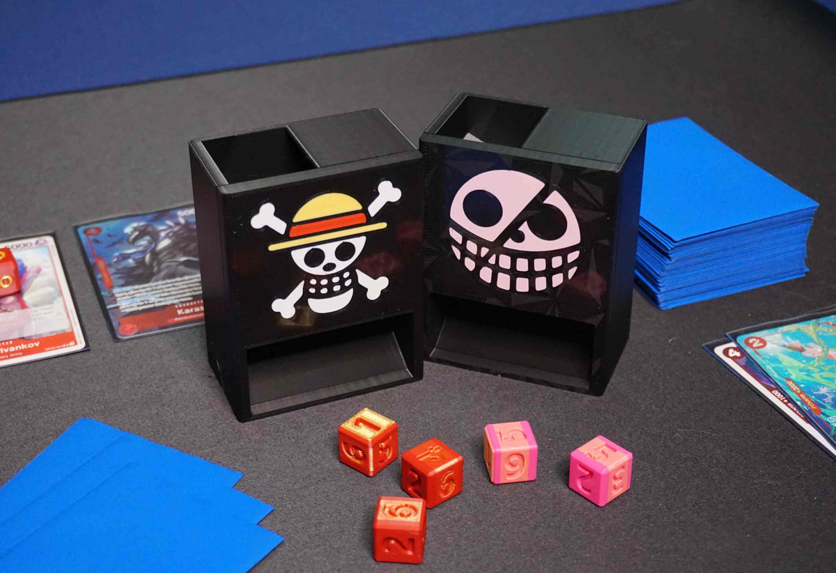 One Piece Mini Dice Towers of the Strawhats and Donquixote jolly rogers on a black playmat with red/gold and pink/gold dice next to a Belo Betty OPTCG deck