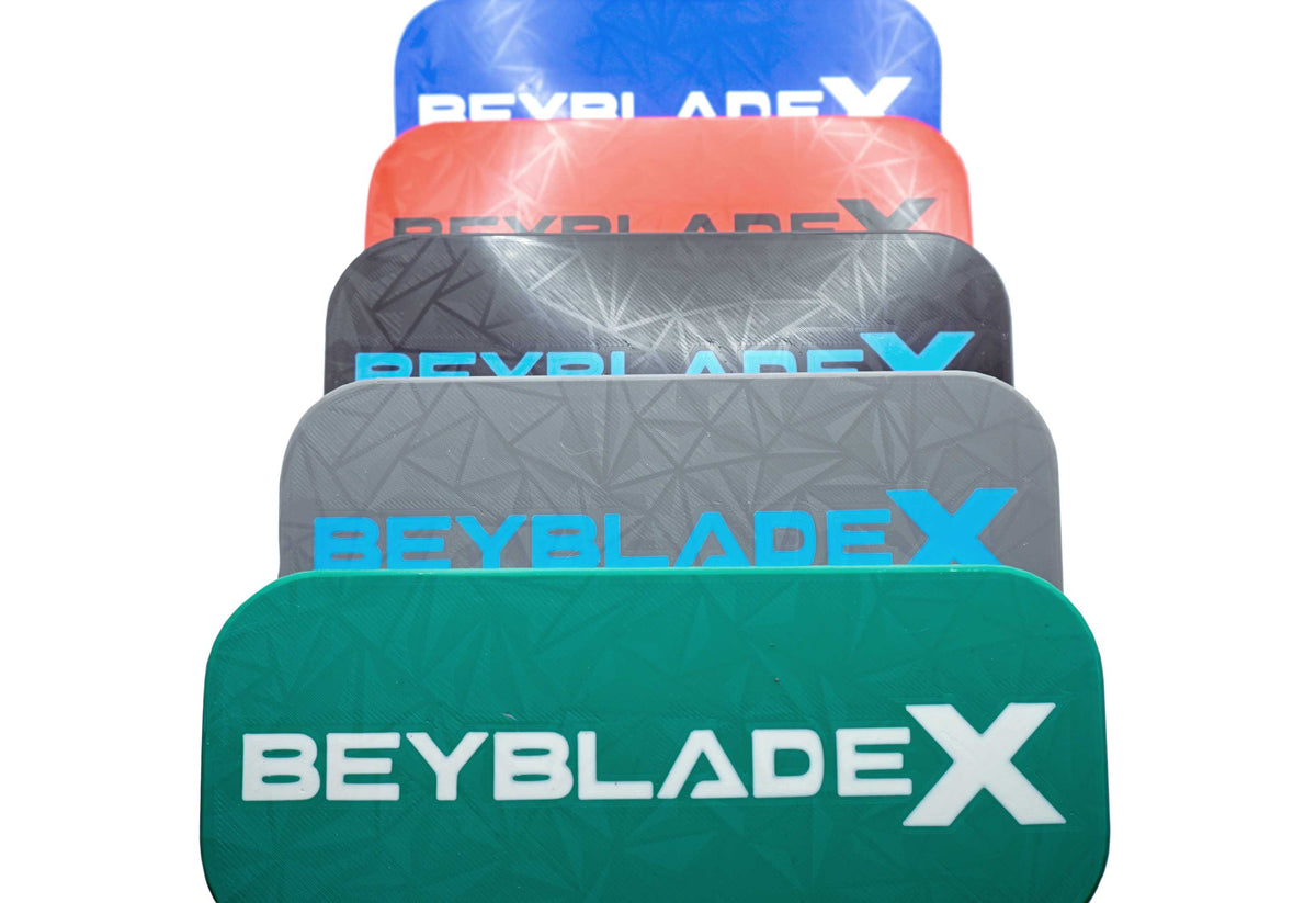 A stack of five Beyblade Pocket deck boxes in shades of green, black, red, gray, and blue. Each deck box features a geometric pattern and the text "BEYBLADE X" in white, black, or sky blue.