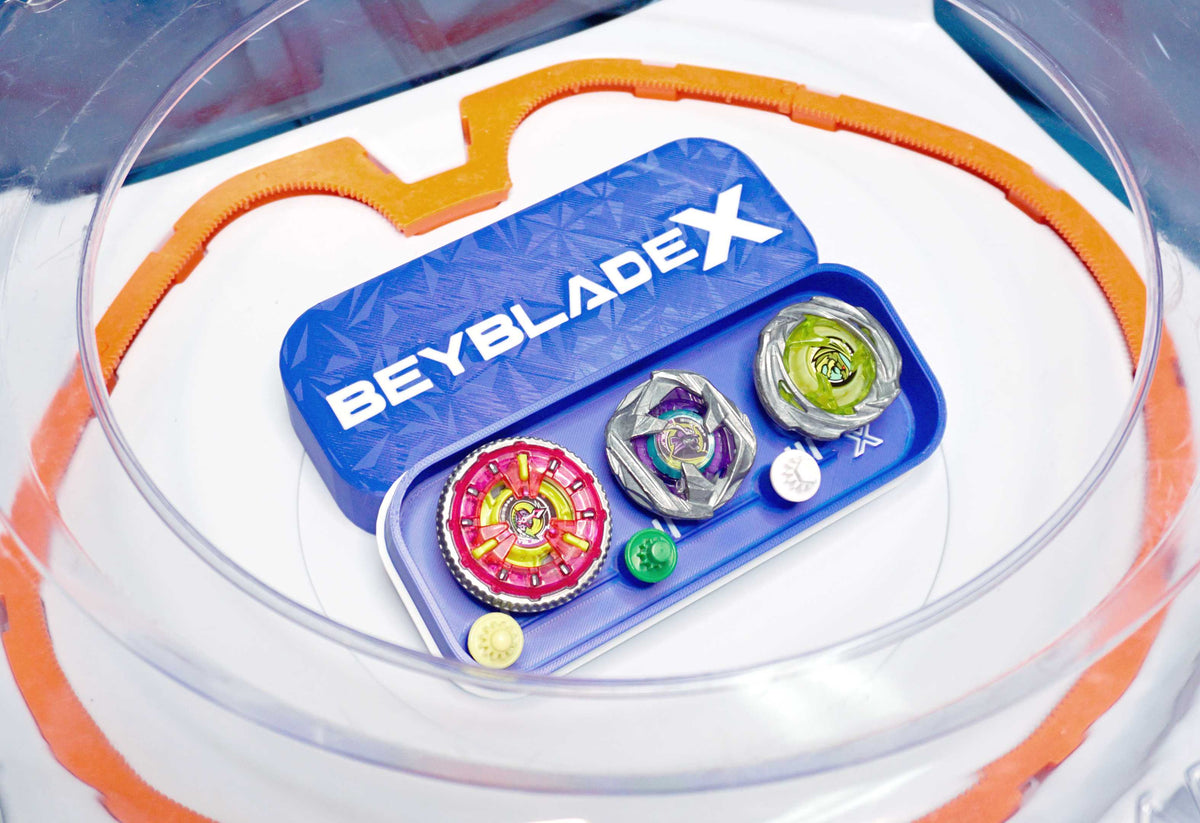 A blue container with the text "BEYBLADE X" holds three colorful spinning tops and small accessories. The set is inside a clear plastic arena with an orange rim.