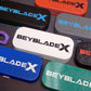 A collection of Beyblade X deck boxes or storage cases in various colors, including black, white, blue, red, gray, and green. Each case features the Beyblade X logo in contrasting colors. Accessories are scattered around the cases.
