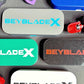 An assortment of rectangular deck cases with the text "BEYBLADE X" in various colors, including gray with blue text, green with white text, and black with red text. Other toy parts are also visible.