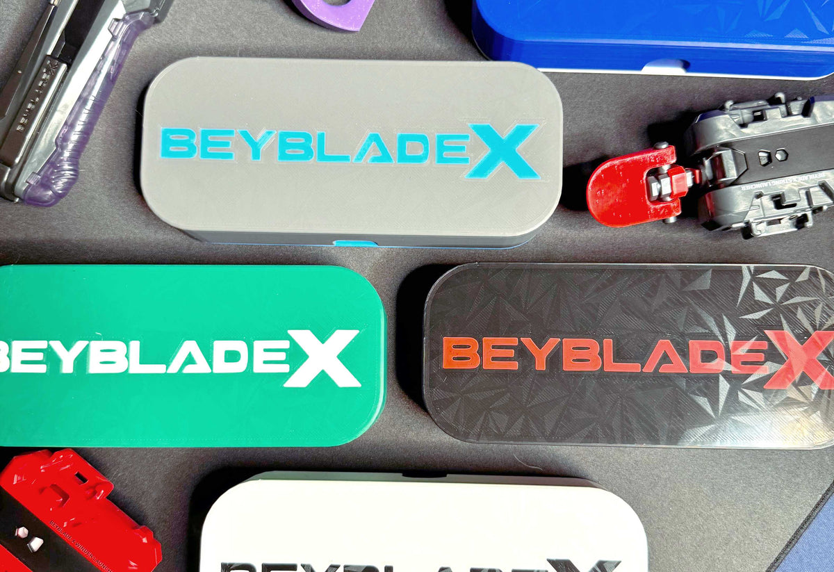 An assortment of rectangular deck cases with the text "BEYBLADE X" in various colors, including gray with blue text, green with white text, and black with red text. Other toy parts are also visible.