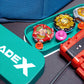 A green container with the text "BEYBLADE X" in white, next to a green tray holding several colorful spinning tops and a red launcher. The tops are various colors, including red, yellow, and silver.