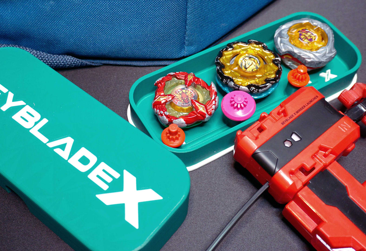 A green container with the text "BEYBLADE X" in white, next to a green tray holding several colorful spinning tops and a red launcher. The tops are various colors, including red, yellow, and silver.
