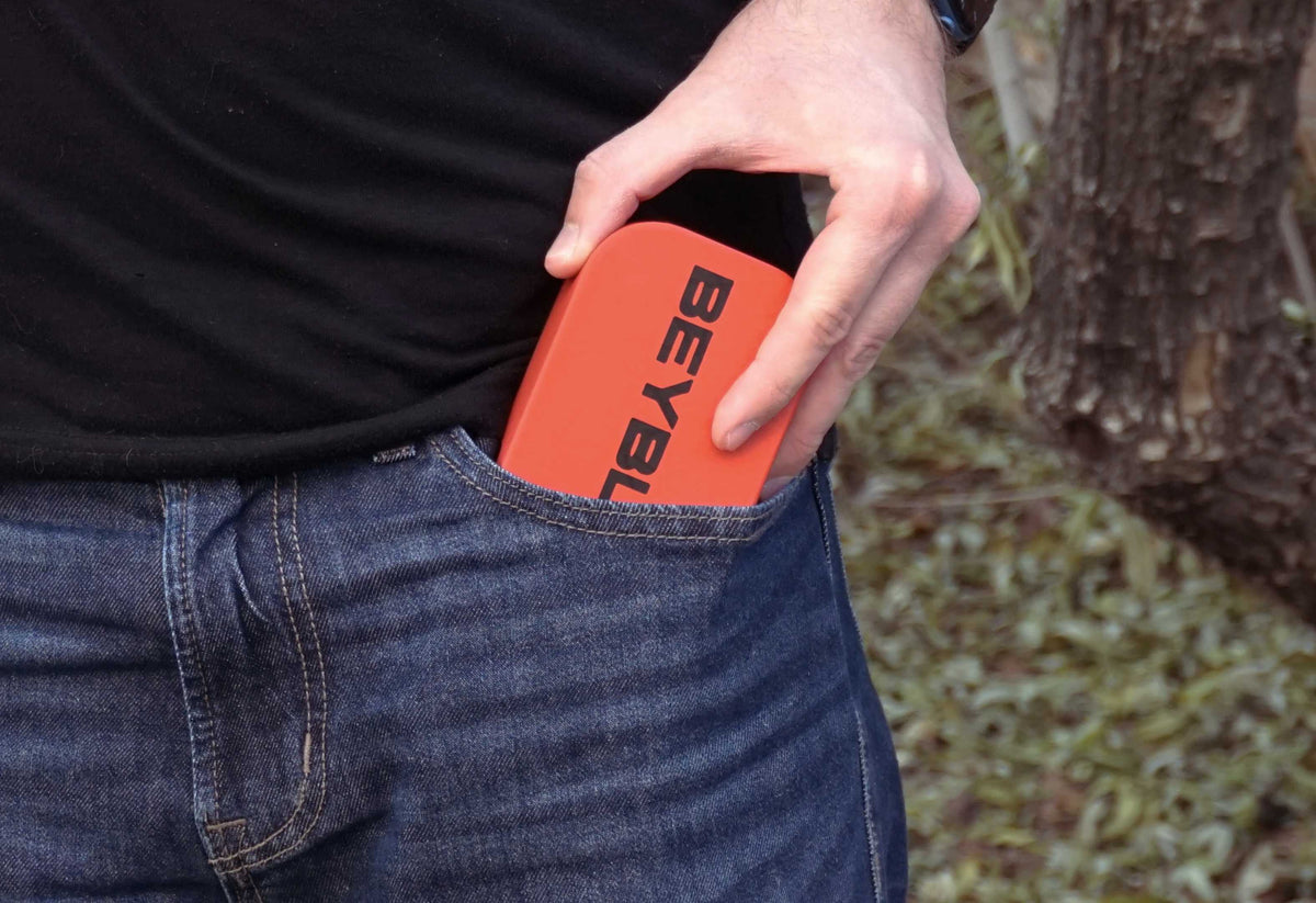 An red Pocket Deck Box with the word "BEYBLADE X" in black is partially visible in a blue jeans pocket. The person is wearing a black t-shirt. The background is blurred.