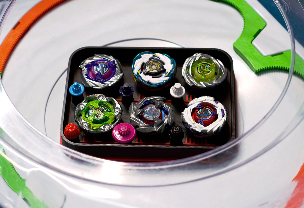 The Pocket Deck Box that holds 6 beys for Beyblade X shown in the black and red color combination. Showcasing Shark Scale, Meteor Dragoon, Wizard Rod, Emperor Pegasus Blast, Cobalt Dragoon, and Hover Wyvern. Sitting inside a Takara Tomy stadium with green and orange X-rails. 